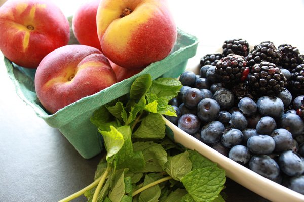 Peaches in a green carton with blueberries and blackberries in a white bowl with sprigs of mint on the side.