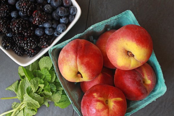 Overhead view of peaches in a green carton with blueberries and blackberries in a white bowl with sprigs of mint on the side.