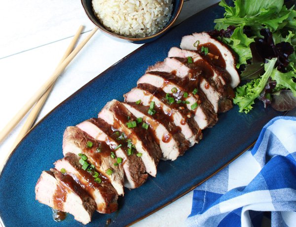 sliced pork tenderloin topped with sauce on a blue serving plate with a side of brown rice and wooden chopsticks