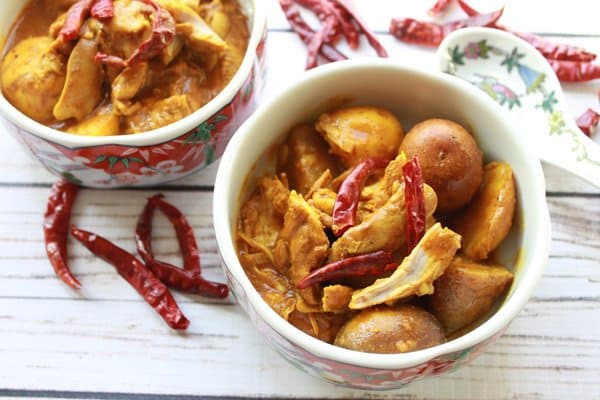 Two bowls filled with Chinese curry chicken and potatoes with pieces of red peppers on top all placed on a white wooden board.