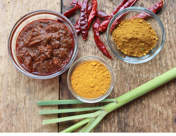 Ingredients in three different glass bowls for making Chinese curry chicken with a lemongrass stalk and red chili peppers on the side on top of a wooden board.