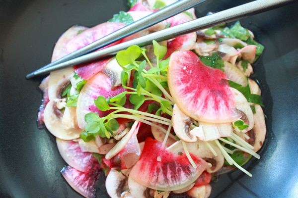 A vibrant mushroom and radish salad topped with sprouts inside a black bowl, with silver chopsticks on the side.