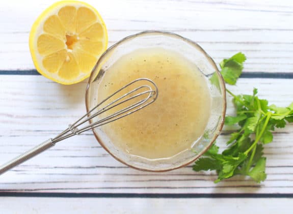 a clear glass bowl with yuzu vinaigrette and a lemon on the side, small whisk and fresh cilantro on a white board.