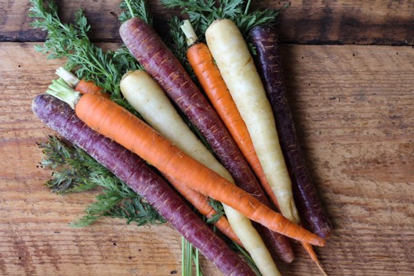 A bunch of raw rainbow carrots with green tops on top of a wooden cutting board.