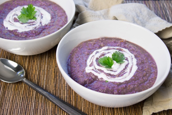 Purple potato and cauliflower soup in two white bowls with a topping of sour cream and garnished with sprigs of cilantro with a spoon and beige napkin on the side on top of a bamboo placemat.