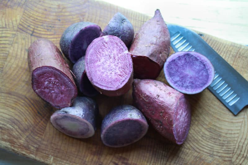 Purple potatos sliced in half with a knife on top of a wooden cutting board.