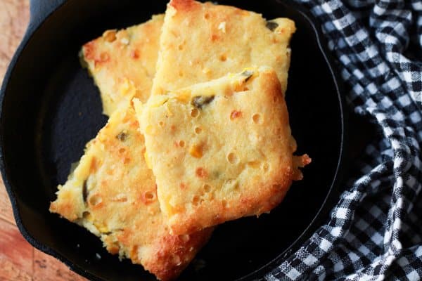 jalapeno cornbread Squares of jalapeño cornbread in a small cast iron skillet on top of a wooden board with a checkered napkin on the side.