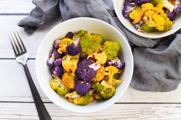 roasted cauliflower Tri-Colored cauliflower florets in two white bowls with a fork and grey napkin on the side on top pf a white plank wooden board.
