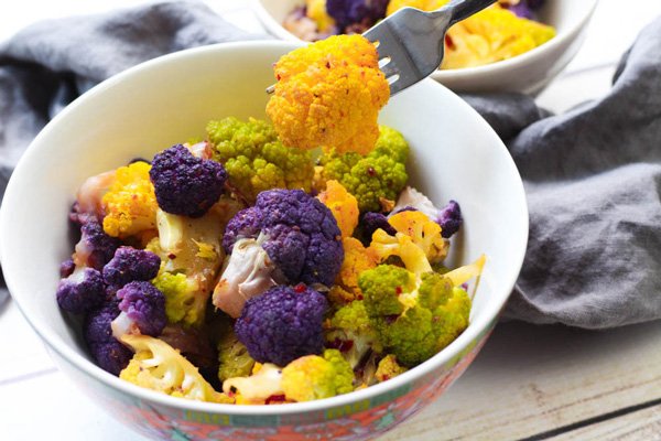 tri-colored cauliflower Tri-colored cauliflower florets in a white bowl with an orange piece of cauliflower held on a fork with a grey napkin on the side on top of a wooden board.