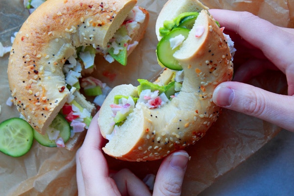 bagel sandwich Two hands holding a California Everything Bagel Sandwich cut in half on top of brown parchment paper with a few slices of cucumber on the side.