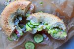 A California Everything Bagel Sandwich cut in half on top of brown parchment paper with a few slices of cucumber on the side.