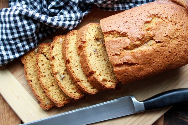 Pumpkin bread A baked loaf of pumpkin bread sliced half-way through on top of a wooden cutting board with a bread knife and black and white checkered napkin on the side.