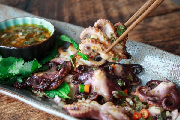 grilled baby octopus Grilled baby octopus on a long gray serving platter with a small bowl of dipping sauce on the side and a pair of chopsticks lifting up a baby octopus.