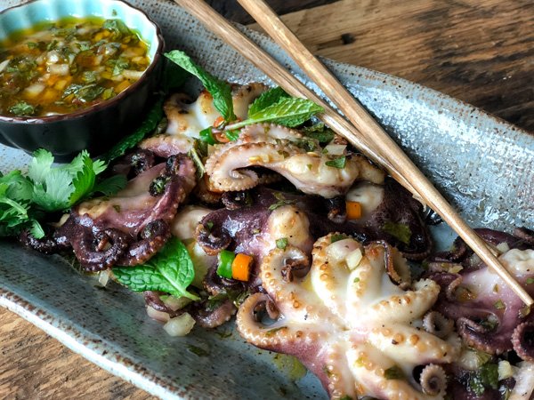 Grilled baby octopus on a long gray serving platter with a small bowl of dipping sauce on the side and a pair of chopsticks.
