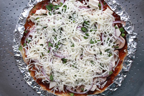 Korean BBQ Cauliflower Pizza Korean BBQ Cauliflower Pizza pie ready to be put in the oven topped with lots of shredded cheese with the edges wrapped with tin foil on top of a silver pizza tray.