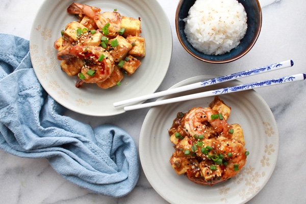 Two white bowls of General Tso's Shrimp and Tofu with chopsticks and a bowls of white rice on the side along with a blue napkin.