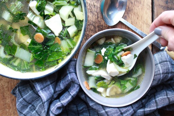 a bowl of Thai Clear Soup with a woman spooning out a spoonful and the pot of soup and a ladle in the background