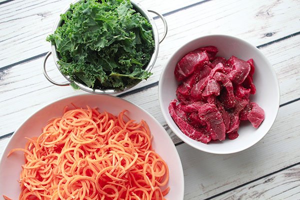 meat, kale, and sweet potatoes Three bowls filled with fresh green kale leaves, sliced sirloin beef, and spiralized sweet potatoes on top of a white plank board.