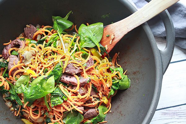 Korean sweet potato noodles with beef Korean Sweet Potato Noodles with Beef being stir-fried in a large wok with a wooden spatula inserted on top of a white plank board.