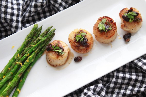 seared scallops with black bean sauce Beautifully plated seared sea scallops drizzled with a black bean sauce with grilled asparagus on the side on top of a long white plate beneath a black and white checkered napkin.