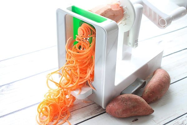 sweet potato noodles A large white spiralizer spiralizing a sweet potato with two whole potatoes on the side on top of a white plank board.