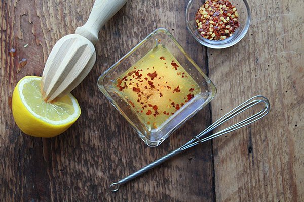 A small square glass bowl filled with a vinaigrette and red pepper flakes with a small bowl of red pepper flakes and a half of a lemon on the side with a small whisk.