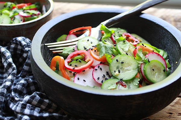 Asian Cucumber Salad A black bowl filled with an Asian cucumber salad with lots of crunchy vegetables and sprinkled with black sesame seeds and topped with a fork with a black and white checkered napkin on the side.