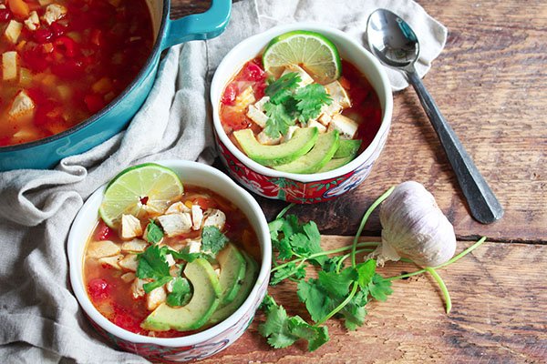 two bowls of chicken tortilla soup on a wooden board with a spoon and veggies along side