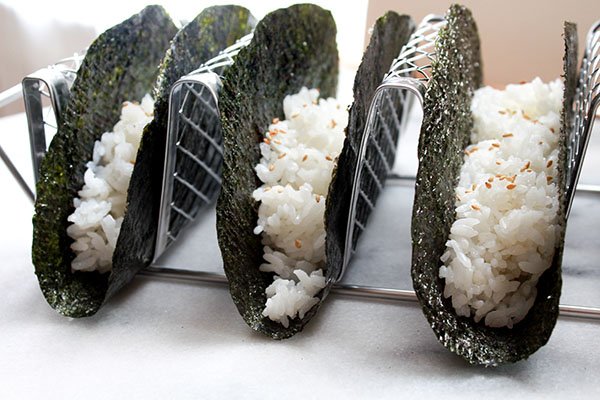 nori and rice Three nori shells in a silver taco holder with white rice nestled inside each shell.