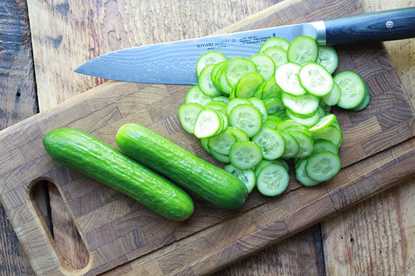 cucumbers Thinly sliced Persian cucumbers in a pile on top of a wooden cutting board with two uncut cucumbers and a chef's knife on the side.