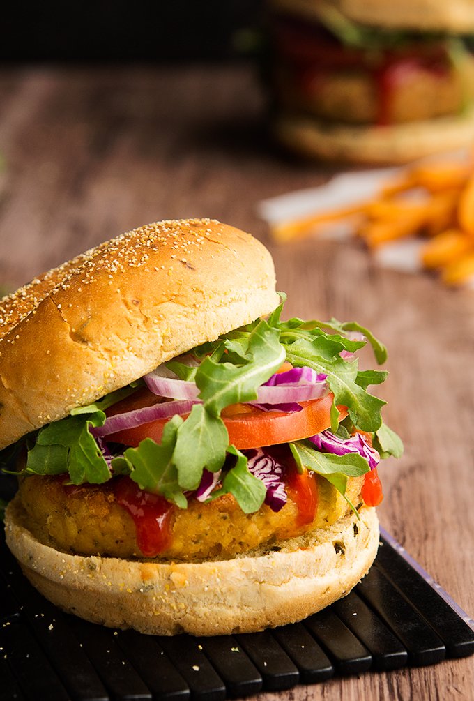 A thick vegetarian tofu burger sandwiched in between a burger bun and topped with greens, tomatoes, and red onions on top of a black plank board with fries on the side.