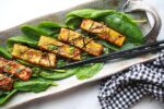 Sesame seared tofu strips on a bed of spinach topped with sesame seeds and seaweed strips presented on a long gray serving plate with black chopsticks on the side.