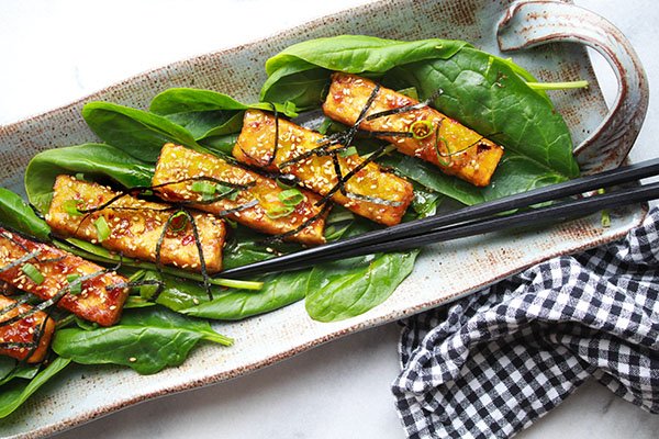 Sesame seared tofu strips on a bed of spinach topped with sesame seeds and seaweed strips presented on a long gray serving plate with black chopsticks on the side.