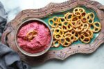 red beet hummus in a bowl with a side of pretzels for dipping on top of a green serving plate