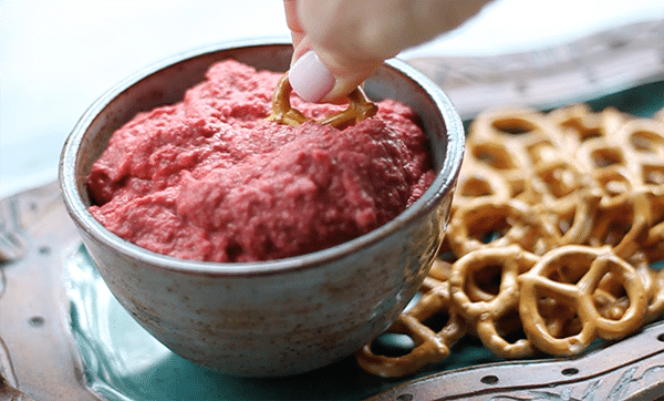 Easy Red Beet Hummus red beet hummus with a pretzel being dipped into a bowl with a side of pretzels on top of a green serving plate