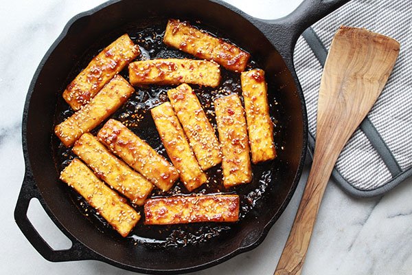 seared tofu strips Golden-brown sesame seared tofu strips searing up in a cast iron skillet with a wooden spatula and oven mitt on the side.
