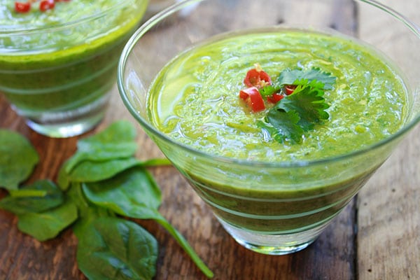 green gazpacho soup in two clear class bowls with fresh spinach on this side on top of a wooden board