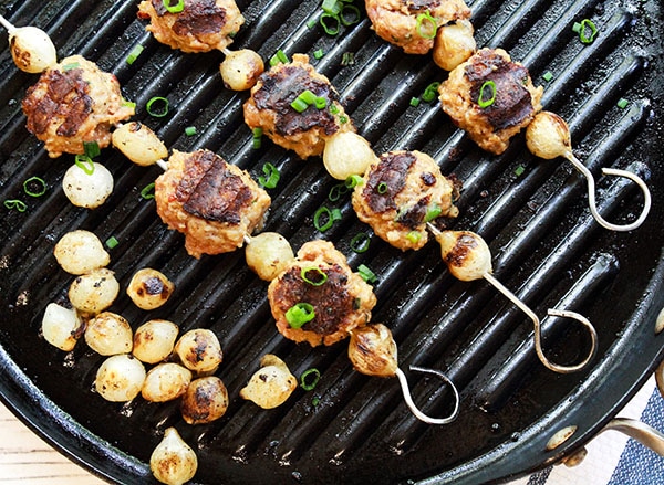 sweet and spicy BBQ meatballs Metal skewers of grilled meatballs with pearl onions in between on top of a grill pan and grilled pearl onions on the side.