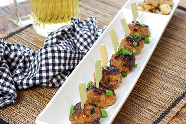 sweet and spicy BBQ meatballs A row of skewered sweet and spicy BBQ meatballs on top of a thin white rectangular serving platter with a black and white checkered napkin and mug of beer on the side.