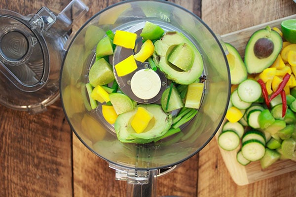 veggies for making gazpacho soup in a food processor with other cut veggies on a wooden cutting board