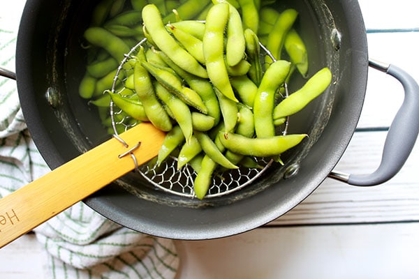 Edamame pods in a strainer on top of a pot of water placed on a white wooden plank board with a kitchen towel on the side.