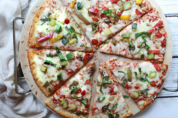 Roasted veggie cauliflower pizza pie cut into slices on top of a pizza stone right out of the oven sitting on a white plank board with a linen napkin on the side.