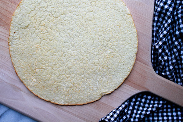 A cauliflower pizza crust right out of the oven on top of a wooden pizza peel with a black and white checkered napkin on the side.