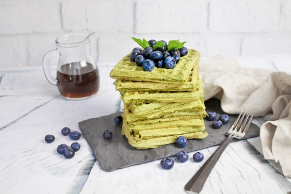 matcha waffles A stack of matcha green tea waffles topped with fresh blueberries placed on top of a black slate tray with a glass pourer filled with maple syrup on the side.
