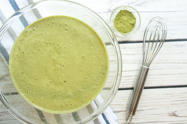 Matcha green tea waffle batter inside a large glass mixing bowl with a whisk and small bowl of match powder on the side on top of a white wooden board.