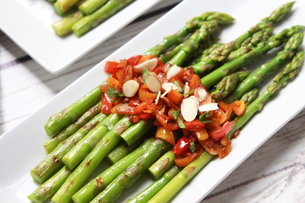 Asian Grilled Sweet and Spicy Asparagus Vibrant grilled asparagus spears on a narrow white plate topped with a roasted pepper and almond sauce.
