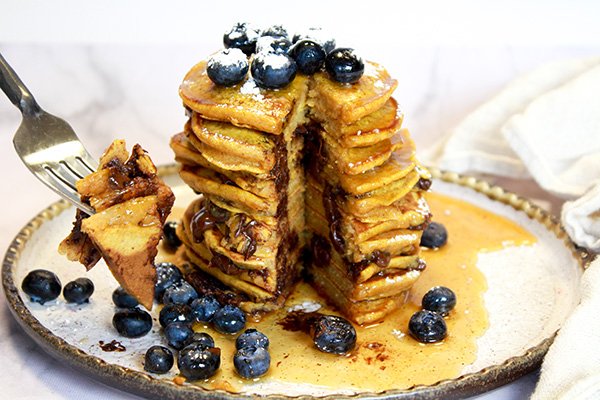 pumpkin five spice pancakes A high stack of pumpkin chocolate chip pancakes topped with fresh blueberries on top of a white plate with a fork and napkin on the side placed on top of a marble surface.