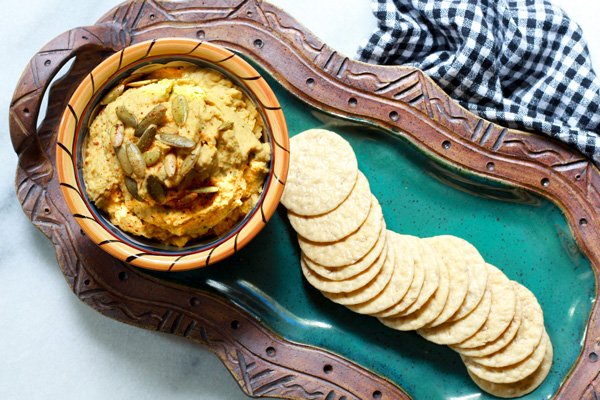 savory pumpkin hummus A small dipping bowl filled with pumpkin hummus topped with pumpkin seeds served on top of a green platter with rice crackers on the side and a black and white checkered napkin on the side.