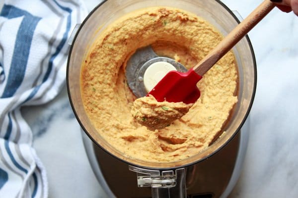 savory pumpkin hummus A food processor with pureed pumpkin hummus inside with a red rubber spatula inserted place on top of a marble surface and a kitchen towel on the side.