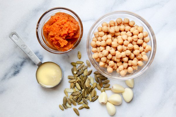 pumpkin hummus Ingredients for making pumpkin hummus placed on a marble surface including chickpeas in a glass bowl, pureed pumpkin in a glass bowl, tahina paste in a small measuring cup and raw pumpkin seeds and garlic cloves.
